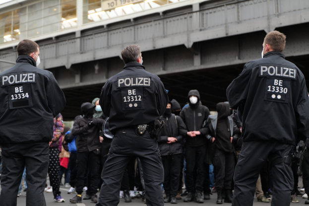 Eine Gruppe von Polizisten in Uniform steht vor einer Menge von Menschen in schwarzen Uniformen und Masken, mit einer Brücke und einem Gebäude im Hintergrund, was auf eine städtische Umgebung hindeutet.