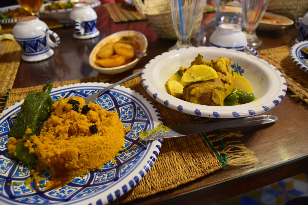 Ein Tisch mit Tellern, Gläsern, Schalen, Krügen, Besteck und Matten bei einem traditionellen marokkanischen Essen, im Hintergrund sitzen Menschen.