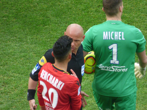 Zwei Fußballspieler und ein Schiedsrichter auf einem grünen Feld, wobei einer einen Ball hält.