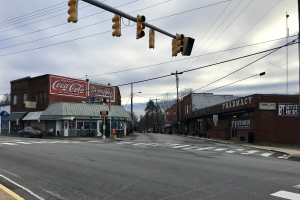 Stadtstraßenkreuzung mit einer Ampel, fahrende Fahrzeuge, Gebäude mit Beschriftung, Strommasten und -leitungen, Bäume und ein bewölkter Himmel.
