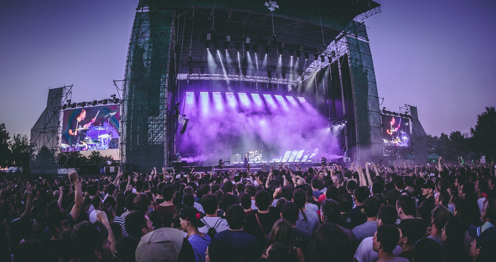 Eine große Menschenmenge auf einem Musikfestival schaut Künstlern auf einer hell erleuchteten Bühne mit Bildschirmen zu, die vor einem Gebäude, Bäumen und einem klaren blauen Himmel aufgebaut ist.