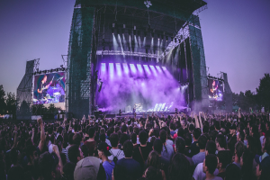 Eine große Menschenmenge auf einem Musikfestival schaut Künstlern auf einer hell erleuchteten Bühne mit Bildschirmen zu, die vor einem Gebäude, Bäumen und einem klaren blauen Himmel aufgebaut ist.