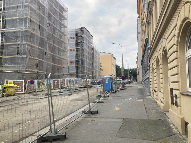 Stadtstraße mit Gebäuden, Fahrzeugen, Straßeninfrastruktur, Bäumen und einer Baustelle mit Gerüsten im Vordergrund unter einem bewölkten Himmel.