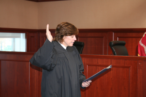 Eine Frau in einer schwarzen Robe steht vor einem Richtertisch, hält eine Akte, mit einem Mikrofon und einer Flagge auf dem Tisch, Stühle dahinter und eine fensterumrandete Wand im Hintergrund.
