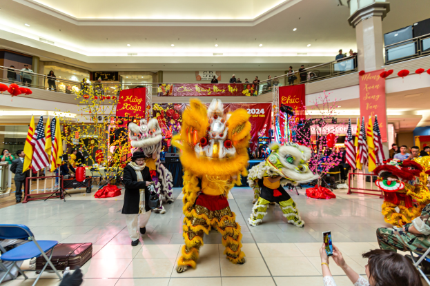 Ein lebendiges chinesisches Neujahrsfest in einem Einkaufszentrum mit Menschen in Kostümen, die tanzen, anderen, die auf Stühlen sitzen und Handys halten, sowie Flaggen, Bannern, Dekorationen, Geländern, Säulen, Deckenleuchten und einem Wasserzeichen unten.
