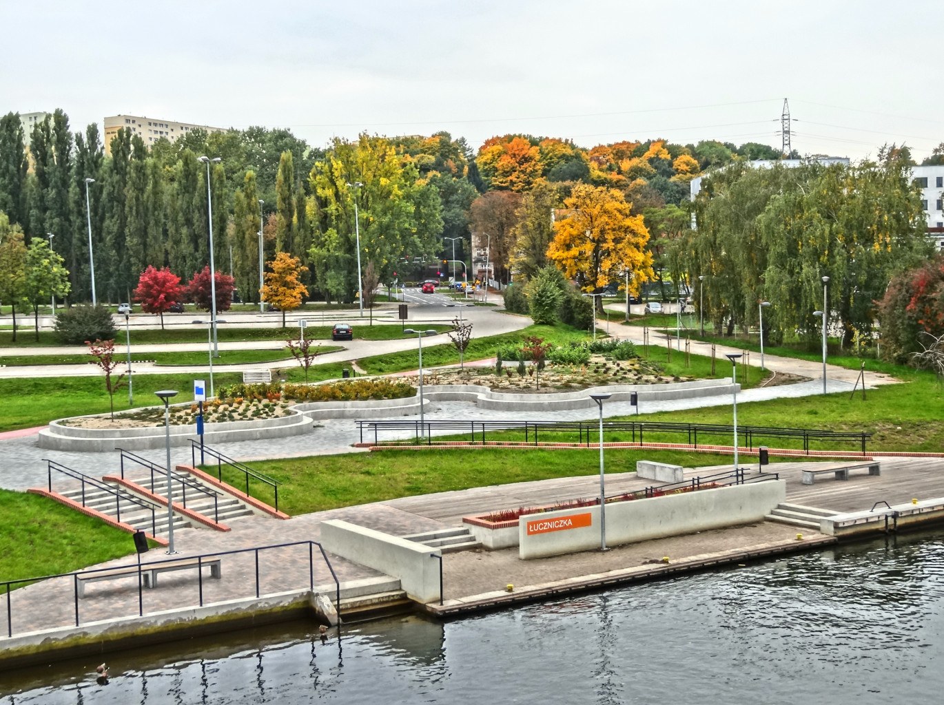 Ein Fluss fließt durch einen grünen Park, der von Bäumen und Gebäuden gesäumt ist, mit Fahrzeugen auf der Straße, Straßenlaternen und Stromleitungen unter einem bewölkten Himmel.