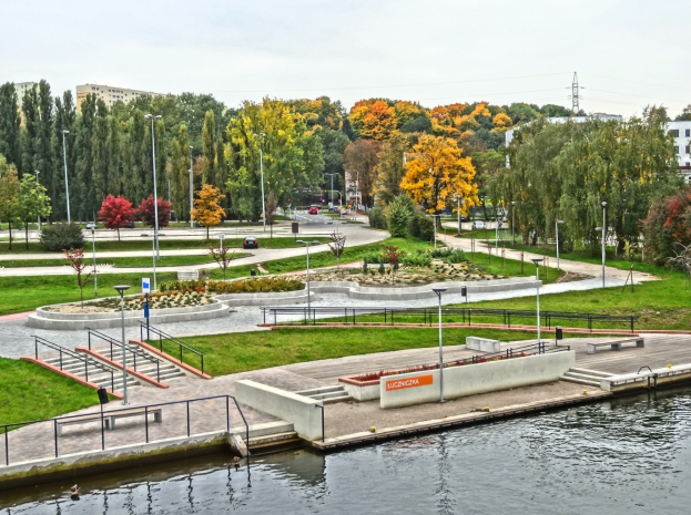 Ein Fluss fließt durch einen grünen Park, der von Bäumen und Gebäuden gesäumt ist, mit Fahrzeugen auf der Straße, Straßenlaternen und Stromleitungen unter einem bewölkten Himmel.