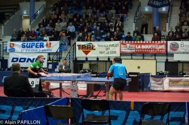 Indoor-Tischtennis-Match mit zwei Spielern, die gegeneinander antreten, sowie einigen stehenden Zuschauern, einer sitzenden Menge, leeren Stühlen und Werbeplakaten in einer Stadionatmosphäre mit sichtbarem Text in der unteren linken Ecke.