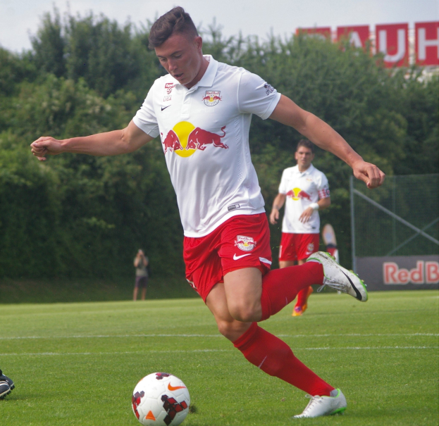 Ein professioneller Fussballspieler tritt einen Ball auf einem grünen Feld mit "RB Leipzig" im Hintergrund.