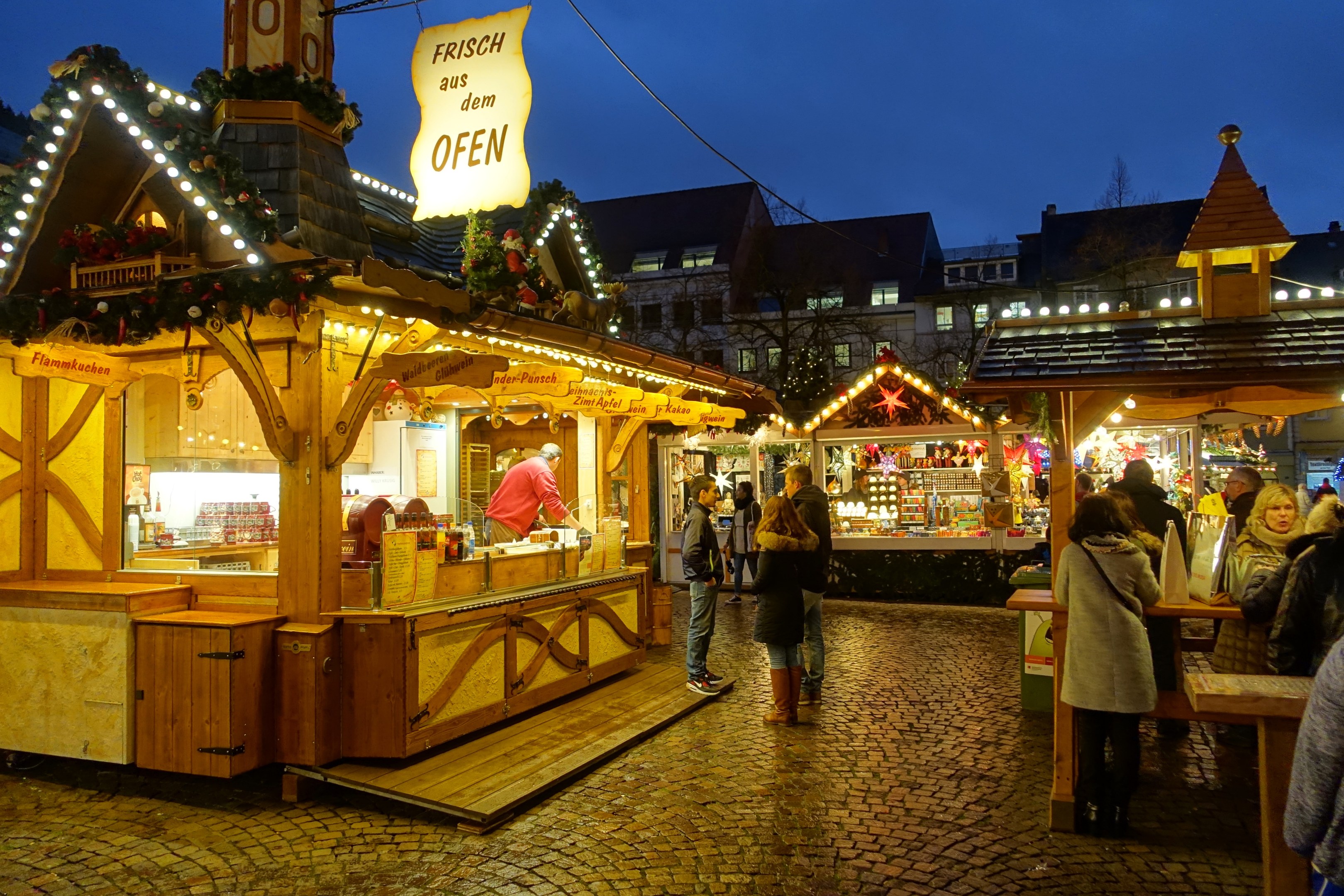 Ein belebter Weihnachtsmarkt auf einer Kopfsteinpflasterstraße bei Nacht, mit Menschen um geschmückte Stände, Gebäuden im Hintergrund, Bäumen und einem klaren Nachthimmel.