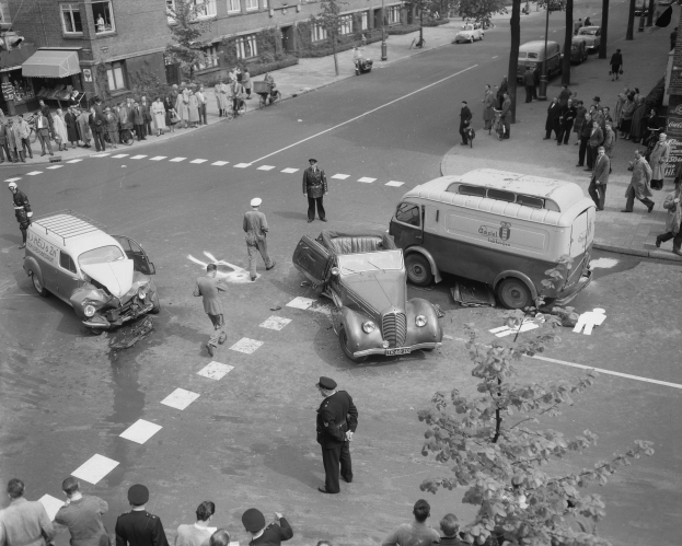 Schwarz-weißes Bild einer Autounfall-Szene an einer Straßenkreuzung mit mehreren Fahrzeugen, darunter ein Lieferwagen, und einer Gruppe von Schaulustigen; Gebäude, Bäume, Pfosten und Texttafeln sind im Hintergrund zu sehen.