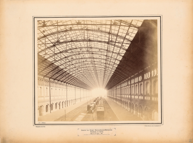 Altes Schwarz-Weiß-Foto eines Bahnhofs mit einer Lokomotive auf den Schienen, flankiert von mehrstöckigen Gebäuden und einem Dach, das von Metallpfosten gestützt wird, mit Text entlang der unteren Kante.