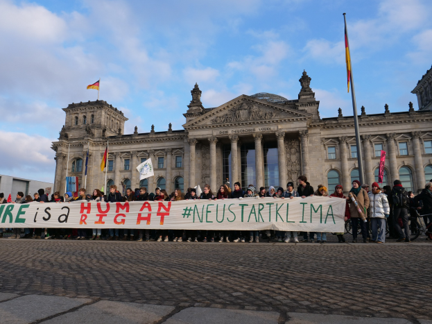 Eine Gruppe von Menschen hält ein Transparent mit der Aufschrift "Wir sind ein Menschenrecht" vor dem Reichstaggebäude in Berlin, Deutschland, mit seinen Säulen, Fenstern, Bögen und Statuen, umgeben von Fahnenmasten und einem bewölkten Himmel.