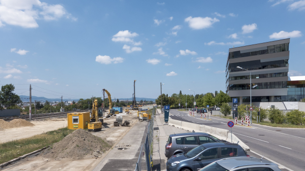 Baustelle mit geparkten Autos, Gebäuden, Straßeninfrastruktur, Vegetation, Sand, Kränen, Bahnschienen, Stromleitungen, Hügel und bewölktem Himmel.