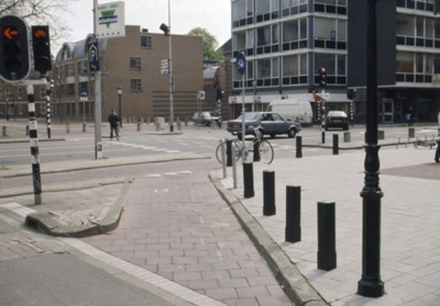 Eine Stadtstraße mit parkenden Autos, Gebäuden, Bäumen, Laternen, Fußgängern auf dem Gehweg und einem Parkautomaten am Boden.