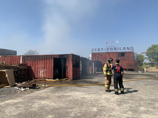 Zwei Feuerwehrleute in Helmen stehen vor einem raucherfüllten Gebäude, einer hält einen Schlauch, mit verstreuter Trümmer, einem Schild, Bäumen und einem klaren Himmel im Hintergrund.