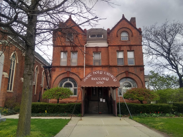 Ein großes Backsteingebäude mit einem Baum davor, umgeben von Gras, Pflanzen, Blumen und Bäumen, unter einem klaren blauen Himmel, mit Fenstern, einer Tür und einer Tafel mit Text, der wahrscheinlich darauf hinweist, dass es der heiligste Friedhof in Detroit ist.