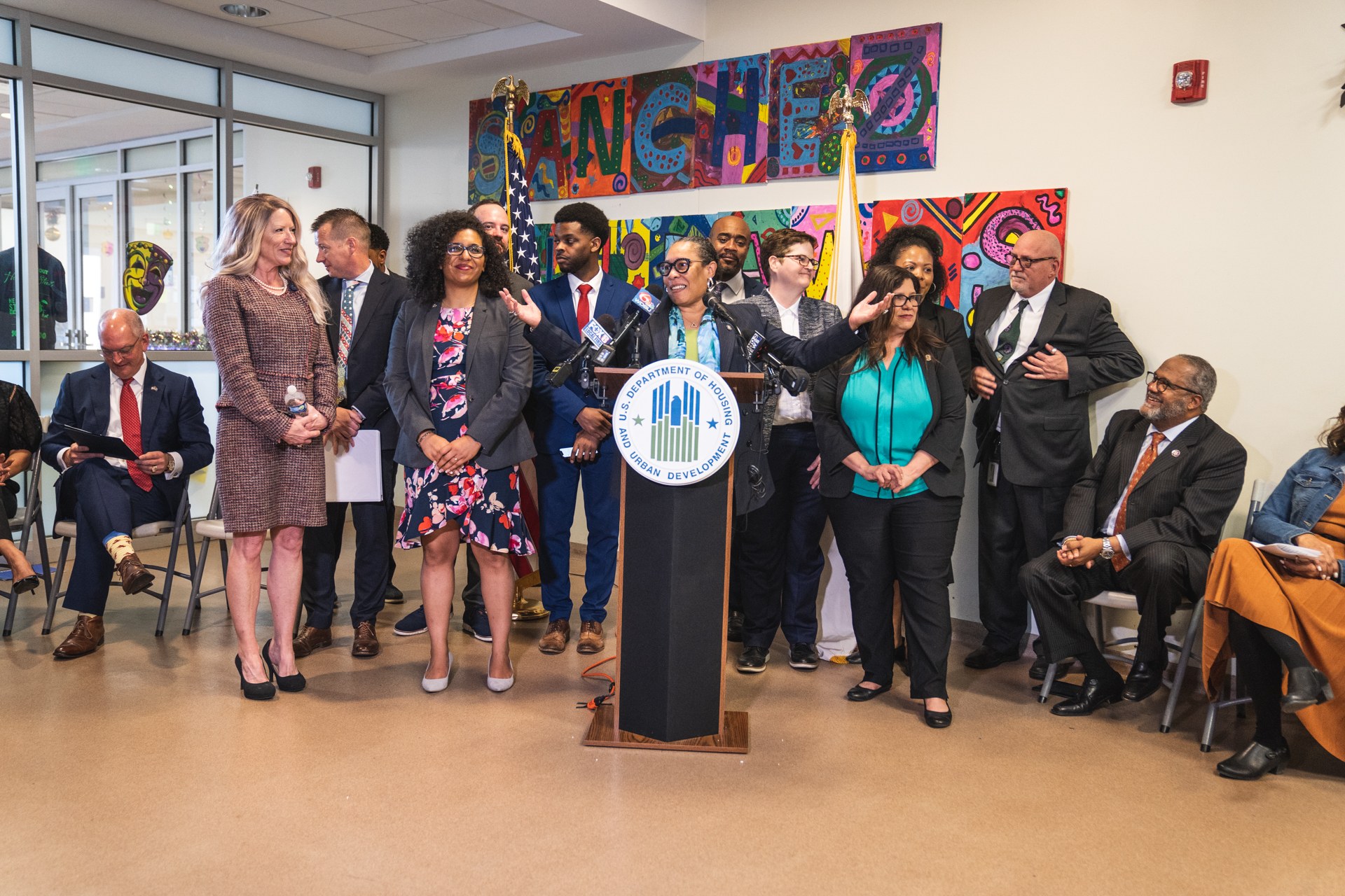 Eine Gruppe von Menschen steht vor einem Podium mit einem Mikrofon und einer Tafel mit Text, während andere auf Stühlen sitzen oder in der Nähe stehen. Flaggen, eine Tafel, ein Feuerlöscher und Deckenleuchten sind an der Rückwand zu sehen.