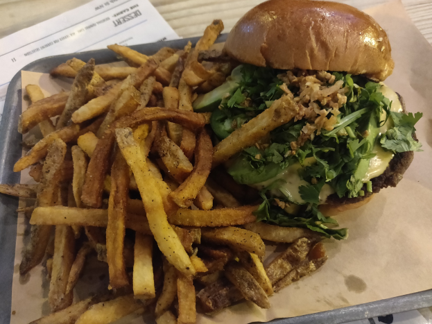 Ein Tablett mit einem Hamburger, der mit Salat, Tomate und Zwiebeln belegt ist, goldenen knusprigen Pommes frites und einem Blatt Papier daneben auf einem Tisch.