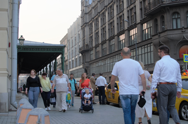 Eine Gruppe von Menschen geht eine Straße entlang, die von hohen Gebäuden gesäumt ist, einige tragen Taschen und einer schiebt einen Kinderwagen, mit Fahrzeugen auf der Straße und städtischen Elementen wie Schildern und Straßenlaternen unter einem bewölkten Himmel.
