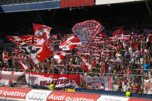 Eine große Menge in einem Stadion hölt Fahnen und Banner, mit einem Metallzaun, einem Schild, einem Anzeigebildschirm und Deckenleuchten.