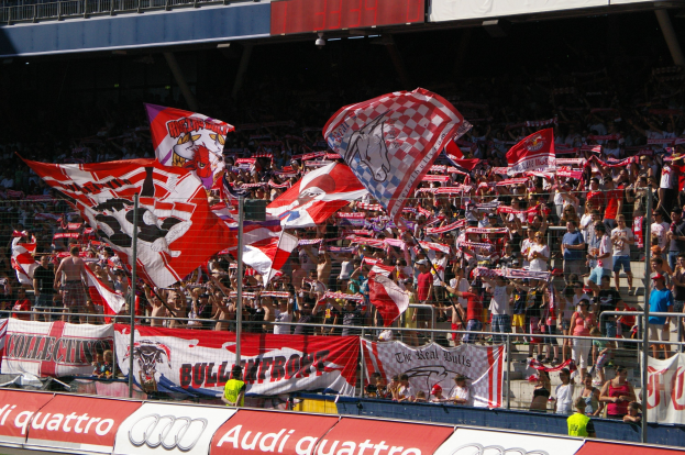 Eine große Menge in einem Stadion hölt Fahnen und Banner, mit einem Metallzaun, einem Schild, einem Anzeigebildschirm und Deckenleuchten.