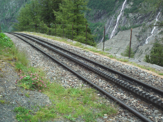 Eine Eisenbahnschiene windet sich durch einen grünen Wald mit einem Wasserfall im Hintergrund, gesäumt von Steinen, Gras und einem Zaun.