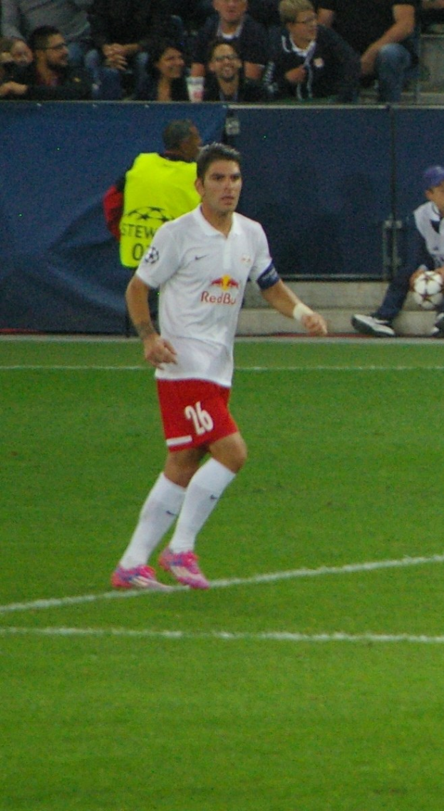 Ein Mann in einem weißen Hemd und roten Shorts, der auf einem Feld Fußball spielt, umgeben von einer Gruppe von Menschen im Hintergrund.