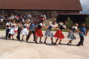 Eine Gruppe von Menschen in traditioneller bayrischer Tracht tanzt vor einem Gebäude mit Dach und Fenstern, einige tragen Hüte und halten Stöcke, während andere auf Bänken sitzen, mit grasbewachsenem Boden und Bäumen im Hintergrund unter einem bewölkten Himmel.