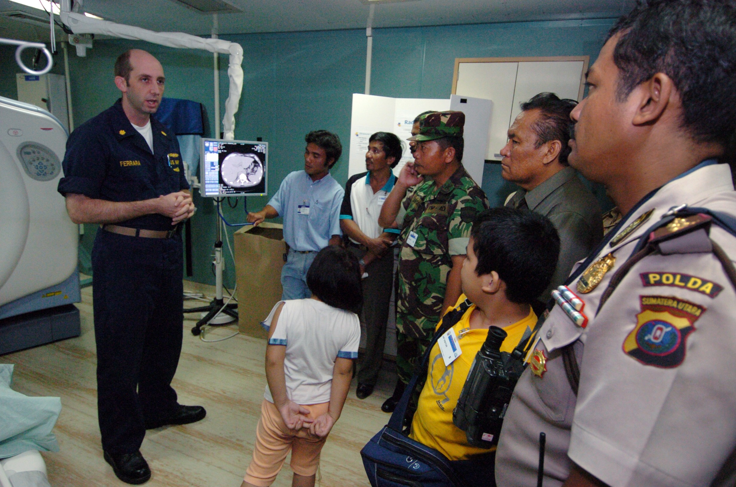 Eine Gruppe von Menschen in einem Krankenhauszimmer mit einem Mann, der ein Mikrofon hält, einem abgedeckten Bett, medizinischen Geräten, einem Röntgenständer, einer Texttafel, einer Tür und Deckenleuchten.