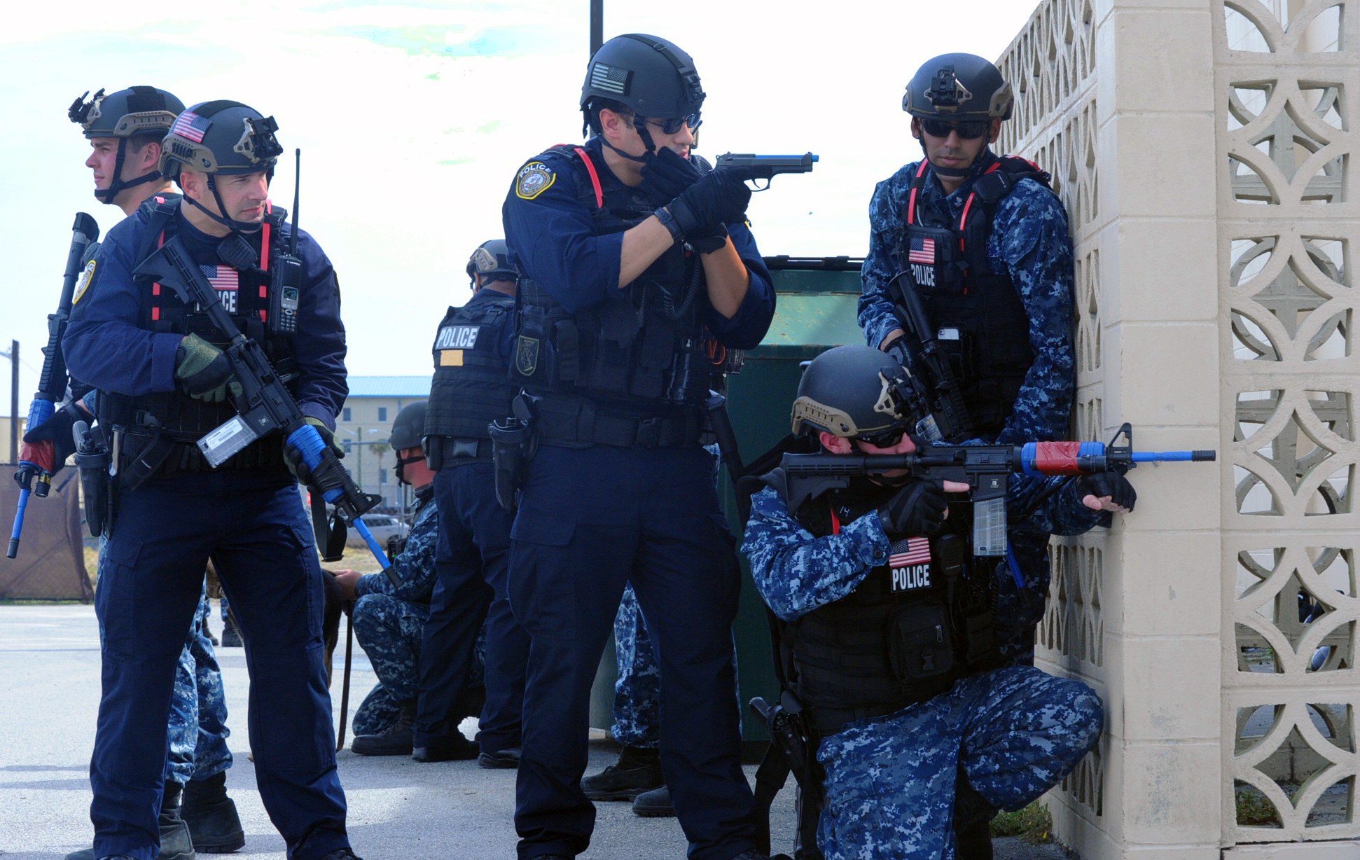 Gruppe von Polizisten in Helmen und taktischer Ausrüstung auf einer Brücke stehend, bewaffnet, mit einer Wand rechts und Gebäuden, Bäumen und Himmel im Hintergrund.