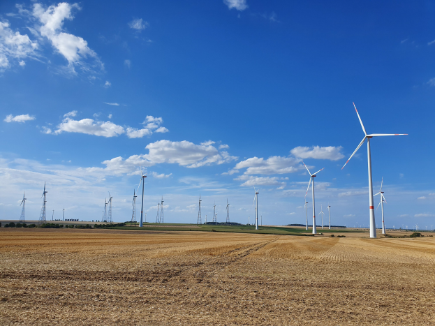 Windräder in einer grünen Wiese mit Bäumen unter einem teilweise bewölkten Himmel.