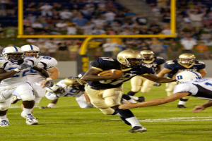 Ein Fussballspieler in Sportuniform und Helm rennt mit dem Ball, umgeben von anderen Spielern, mit unscharfen Hintergrundelementen und Text unten.