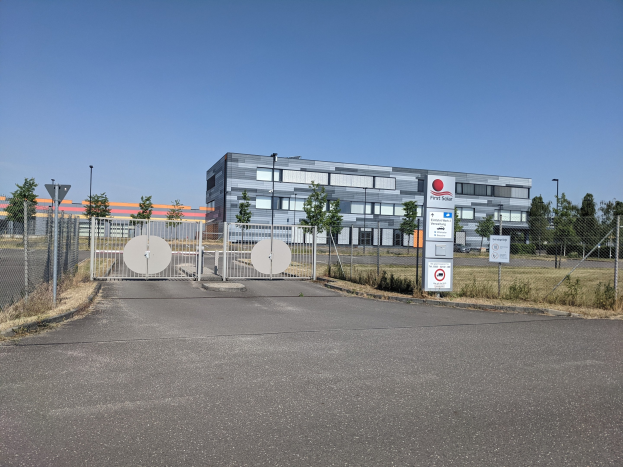 Großes industrielles Gebäude mit der Aufschrift "First Solar" und zahlreichen Fenstern, umgeben von einem Metallzaun, Bäumen, Pflanzen, Gras, Schildern, Straßenlaternen und einem Weg unter einem bewölkten Himmel.