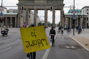 Eine Gruppe von Radfahrern in Helmen fährt eine Straße vor dem Brandenburger Tor in Berlin, Deutschland, entlang, wobei eine Person ein gelbes Schild hält, Lichtmasten, Verkehrszeichen, Gebäude, Bäume und einen klaren blauen Himmel im Hintergrund zu sehen sind.