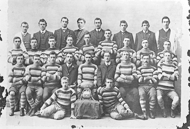 Schwarzes und wei├čes Foto einer Fuballmannschaft mit sitzenden und stehenden Spielern, einem Helm in der Mitte und einem leicht unscharfen Hintergrund.