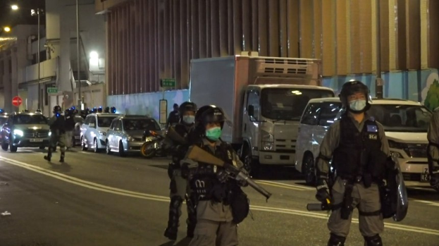 Polizeibeamte in Helmen, Handschuhen und Gesichtsmasken stehen nachts auf einer Straße, einige halten Waffen, mit Fahrzeugen, Laternen, Schildern und Gebäuden im Hintergrund.