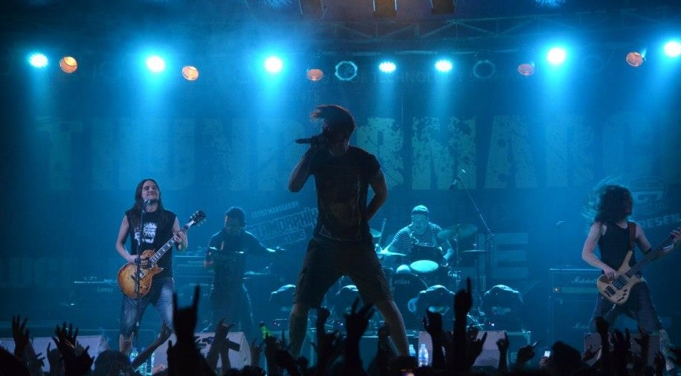 Eine Gruppe von Musikern auf einer hell erleuchteten Bühne mit Gitarren, einem Mikrofon und Trommeln, während die Zuschauer im Vordergrund die Hände heben.