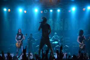 Eine Gruppe von Musikern auf einer hell erleuchteten Bühne mit Gitarren, einem Mikrofon und Trommeln, während die Zuschauer im Vordergrund die Hände heben.