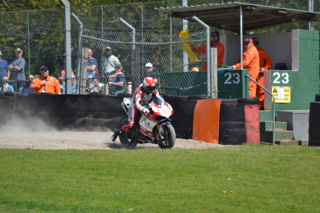 Ein Motorradfahrer mit Helm und Handschuhen fährt über ein grasbewachsenes Feld, mit Zuschauern in Mützen und Schutzbrillen, einem Zaun mit Bäumen, Treppen mit Geländern und einem nummerierten Schuppen mit Texttafeln im Hintergrund.