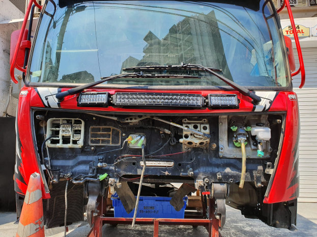Roter Lastwagen vor einem Gebäude mit einem Verkehrskegel auf der linken Seite und einem Rollladen auf der rechten Seite geparkt, mit nummerierten Schildern im Hintergrund, die Wartungsarbeiten anzeigen.