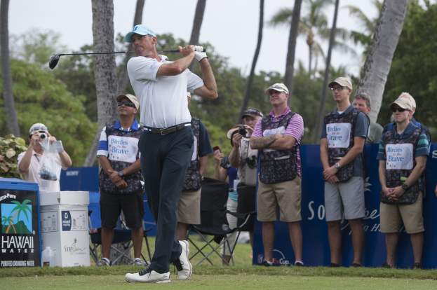 Ein Mann in einem weißen T-Shirt, schwarzen Hosen und einer blauen Mütze schwingt einen Golfschläger auf einem grünen Feld, mit Zuschauern und Kameras hinter ihm unter einem klaren blauen Himmel und Bäumen.