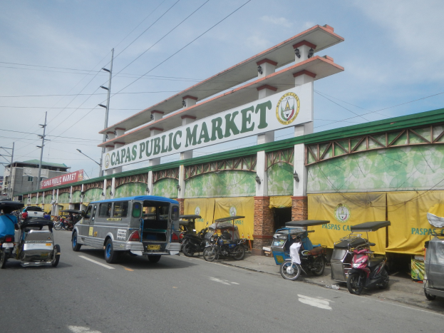Eine belebte Stadtstraße mit Autos, Motorrädern und Rikschas, die an dem Gebäude "Capas Public Market" vorbeifahren, vor einer Kulisse aus Strommasten, Gebäuden und bewölktem Himmel.