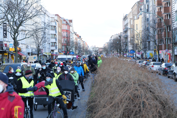 Eine große Gruppe von Menschen in Masken und Sicherheitswesten auf Fahrrädern eine von Bäumen gesäumte Straße entlang mit Gebäuden, Fahrzeugen, Laternenmästen und Texttafeln, unter einem klaren blauen Himmel.
