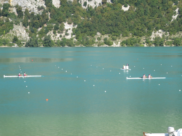 Eine Gruppe von Menschen, die in einem Rennen auf einem Gewässer rudern, mit einem baumbewachsenen Hügel im Hintergrund und einem Boot unten sichtbar.