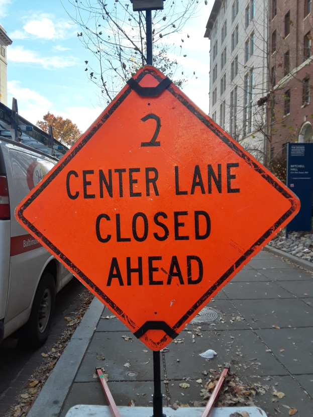 Orangefarbenes Schild mit der Aufschrift "2 Center Lane Closed Ahead" auf einer Straße mit Fahrzeugen, Pfosten, Brettern, Bäumen, Gebäuden und einem bewölkten Himmel im Hintergrund.