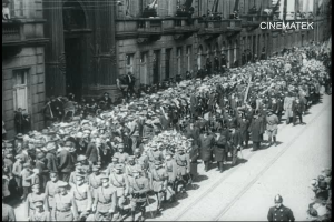 Schwarzes und weißes Foto einer großen Menge, die bei einer Parade marschiert, einige halten Gewehre, vor einem Gebäude mit einem Wasserzeichen in der oberen rechten Ecke.