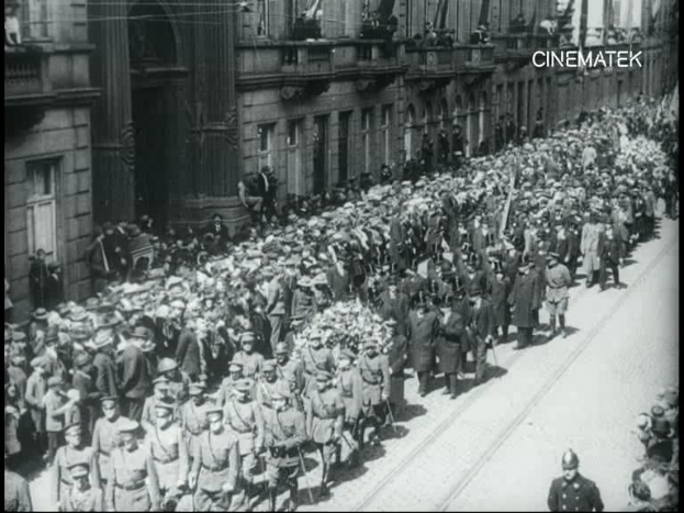 Schwarzes und weißes Foto einer großen Menge, die bei einer Parade marschiert, einige halten Gewehre, vor einem Gebäude mit einem Wasserzeichen in der oberen rechten Ecke.