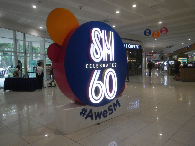 Eine Gruppe von Menschen steht auf einem Einkaufszentrumsboden unter einem "SM feiert 60. Geburtstag"-Schild, mit einem schwarzen bedeckten Tisch auf der linken Seite und Glasdoors, Schildern und Deckenleuchten im Hintergrund.