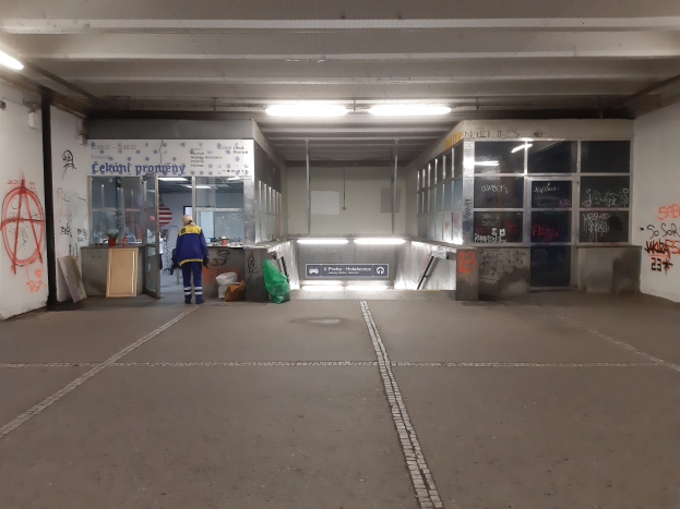 Ein Mann in einer blauen Jacke und Hose steht in einer leeren Parkgarage mit Graffiti an den weißen Wänden und Lampen an der Decke, mit einem Müllcontainer davor.